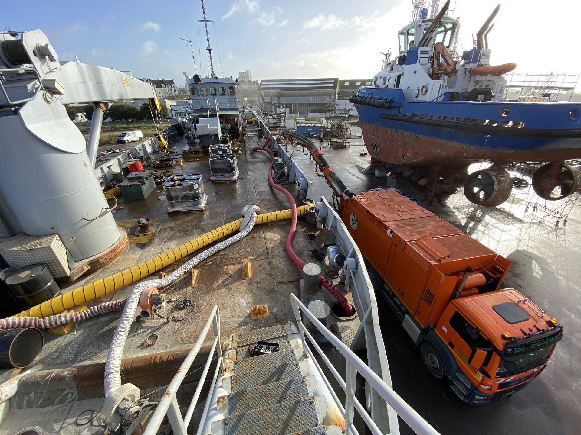 Vider la cale d'un bateau au Port de commerce de Lorient ; une longueur de tuyau supplémentaire est ajoutée à l'embout du bras de l'aspiratrice afin d'accéder aux cales du bateau. Travaux préparatoire au sablage de la cale de bateau.